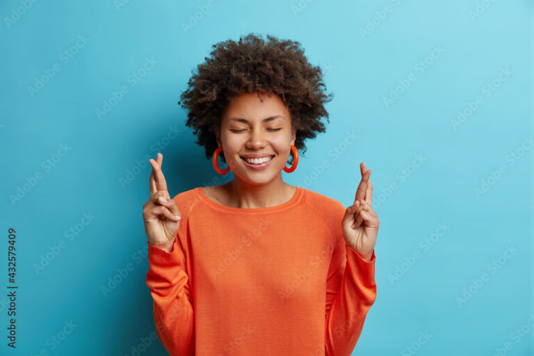 Lovely cheerful young African American woman with toothy perfect smile keeps fingers crossed believes in good luck wears orange jumper isolated over blue backgound. Superstitious female makes wish